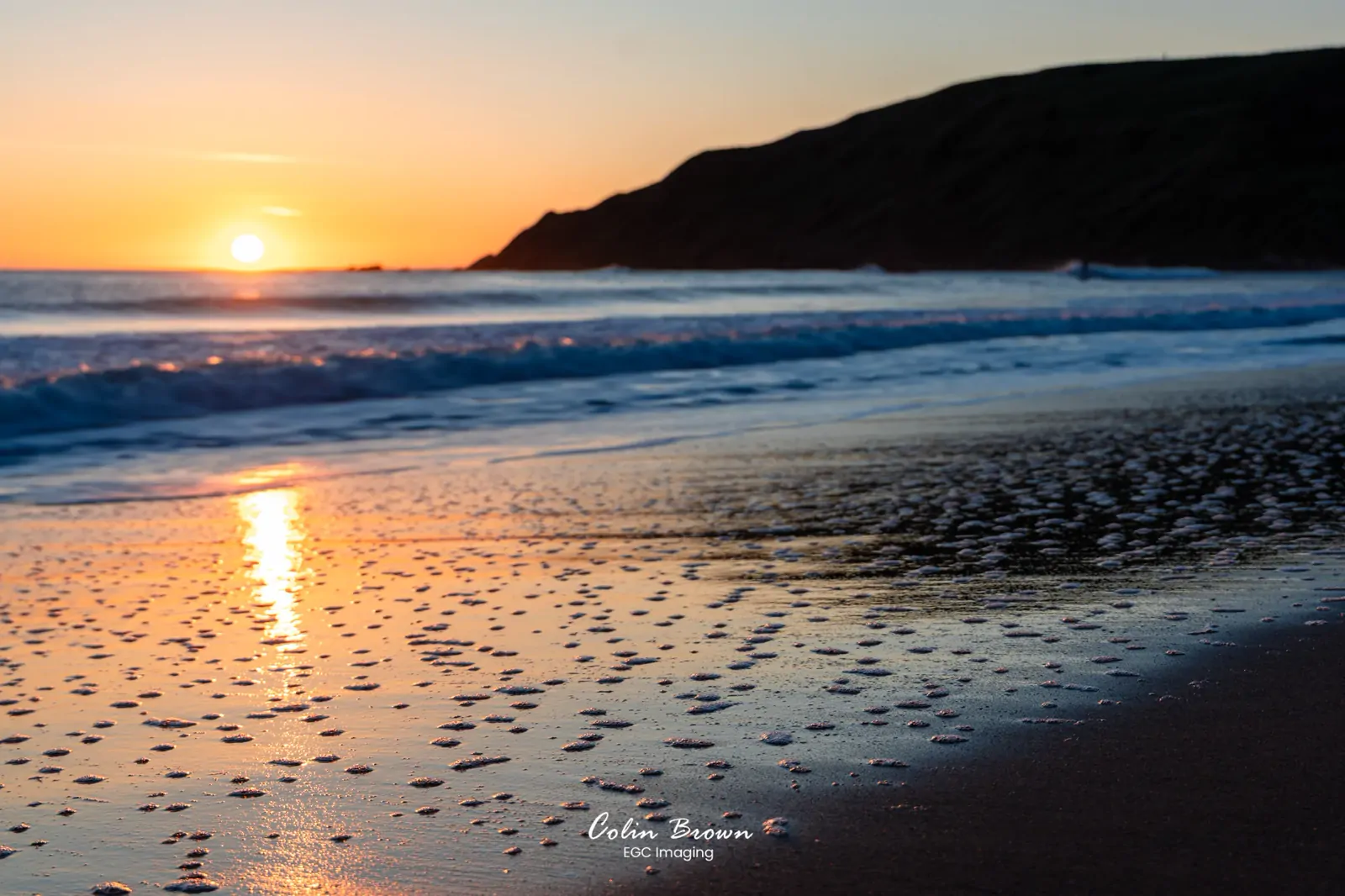 Close-up of sea foam bubbles on wet sand at sunset with a headland behind