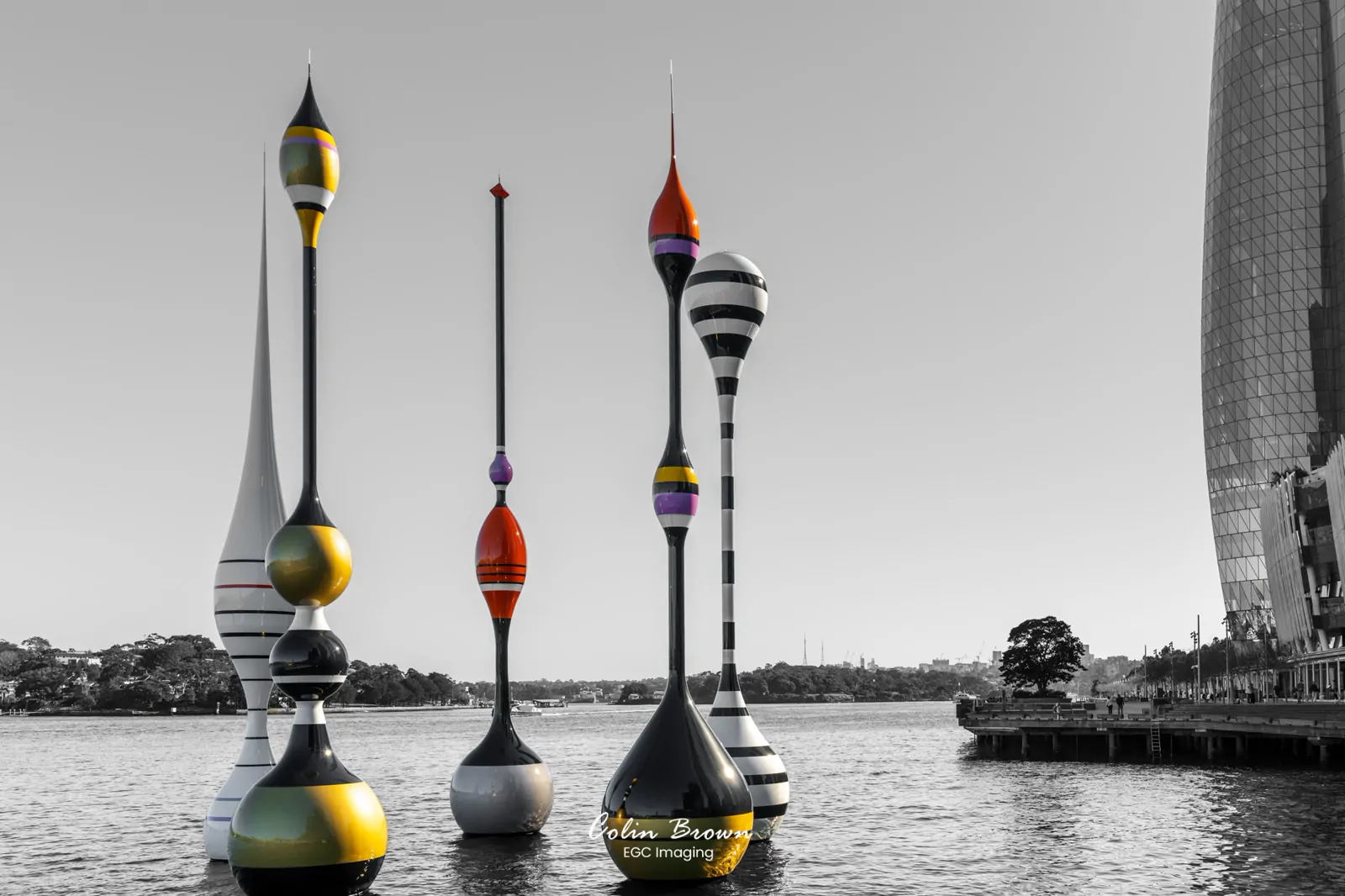 Sculptural buoys on a harbour with a city skyline behind, in selective colour