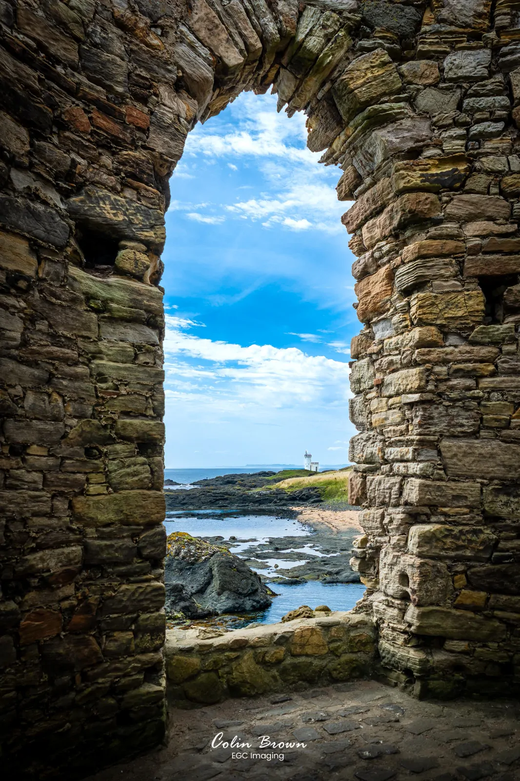 View through a ruined castle window overlooking a lighthouse on the coast