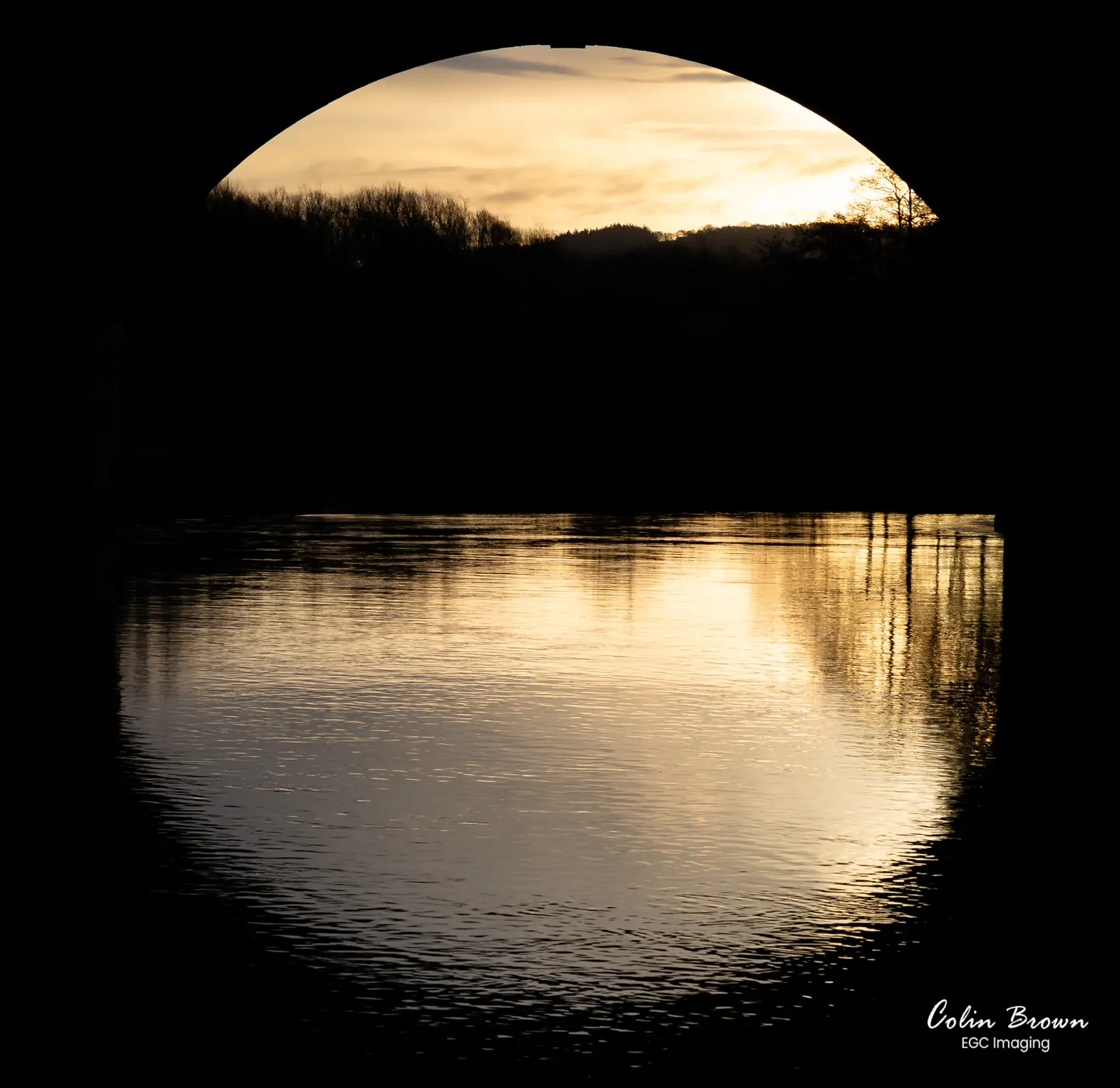 Sunset silhouette viewed through a stone bridge arch over calm water