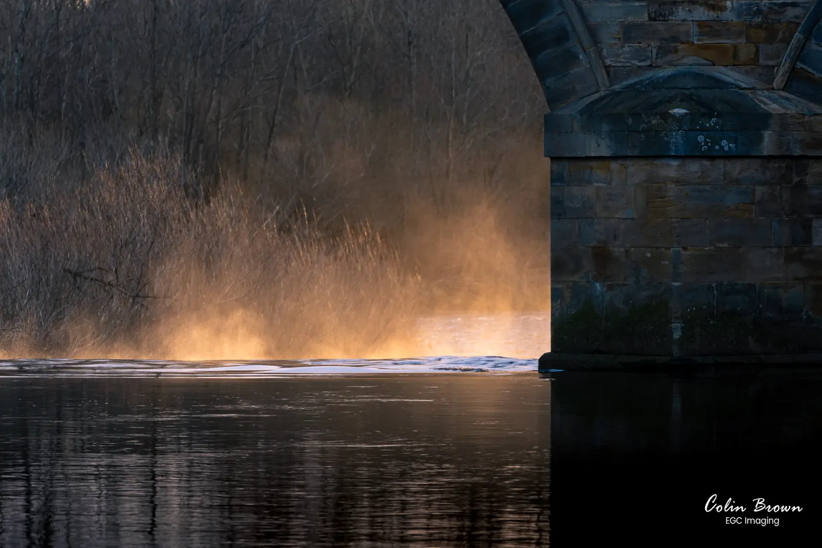 Golden mist rising from a river beneath a stone bridge arch at sunset